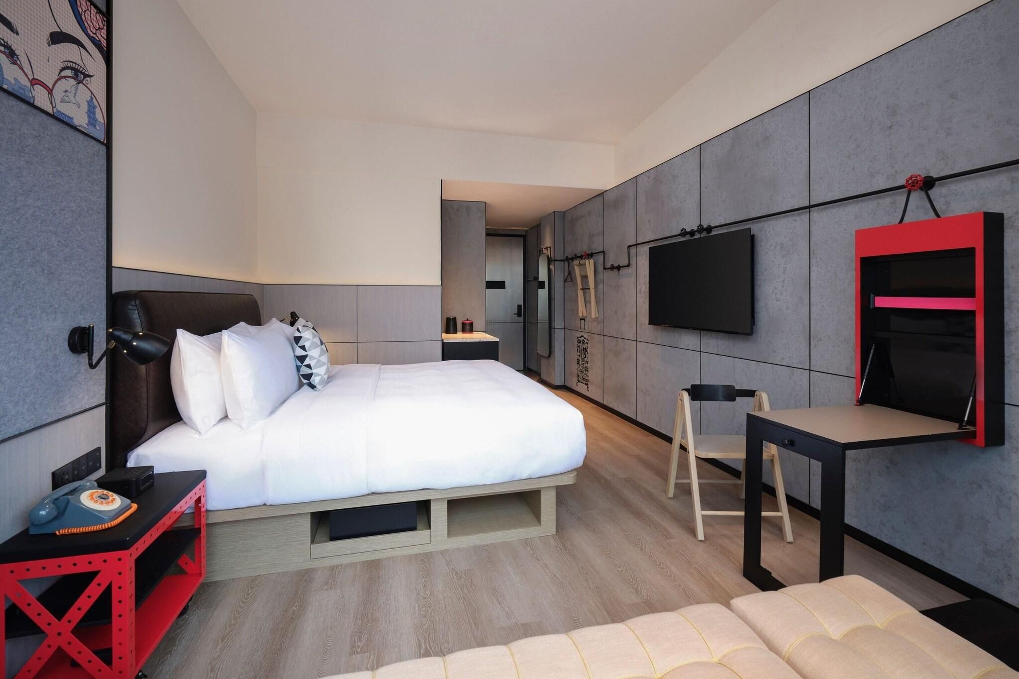 Una habitación de hotel moderna con cama, mesita de noche, silla, televisión y un pequeño escritorio.