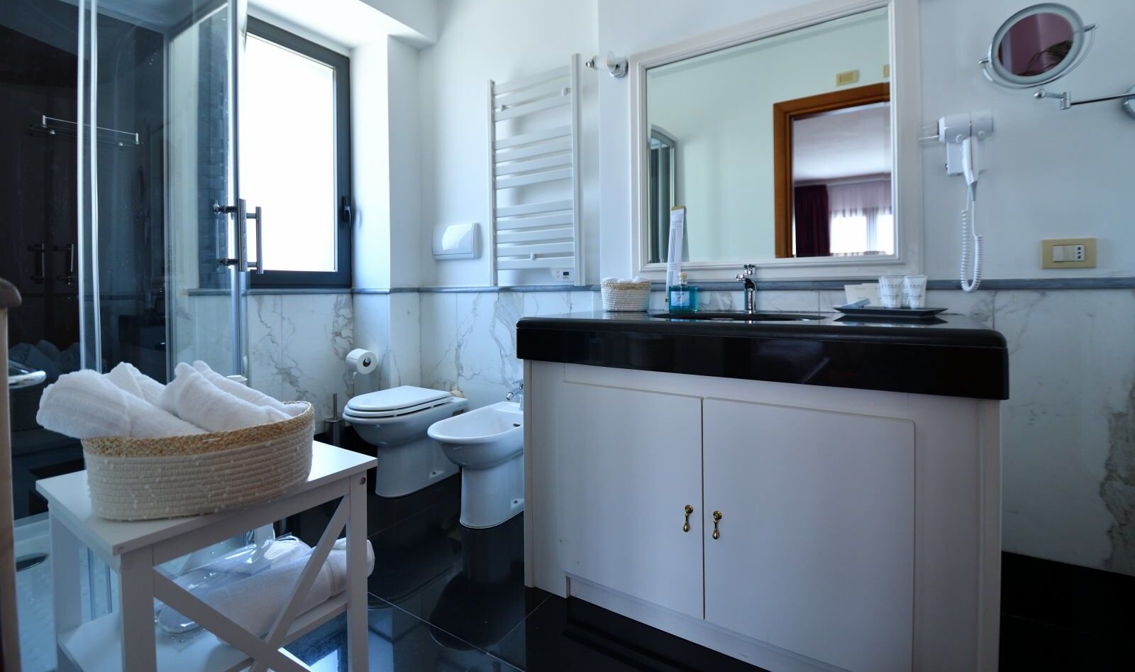 Een badkamer met toilet, bidet en wastafel met spiegel erboven. Er staat een mand handdoeken op een klein tafeltje.