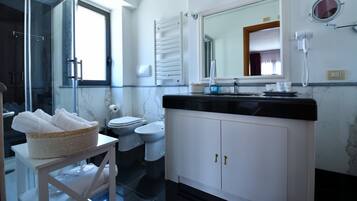Een badkamer met toilet, bidet en wastafel met spiegel erboven. Er staat een mand handdoeken op een klein tafeltje.