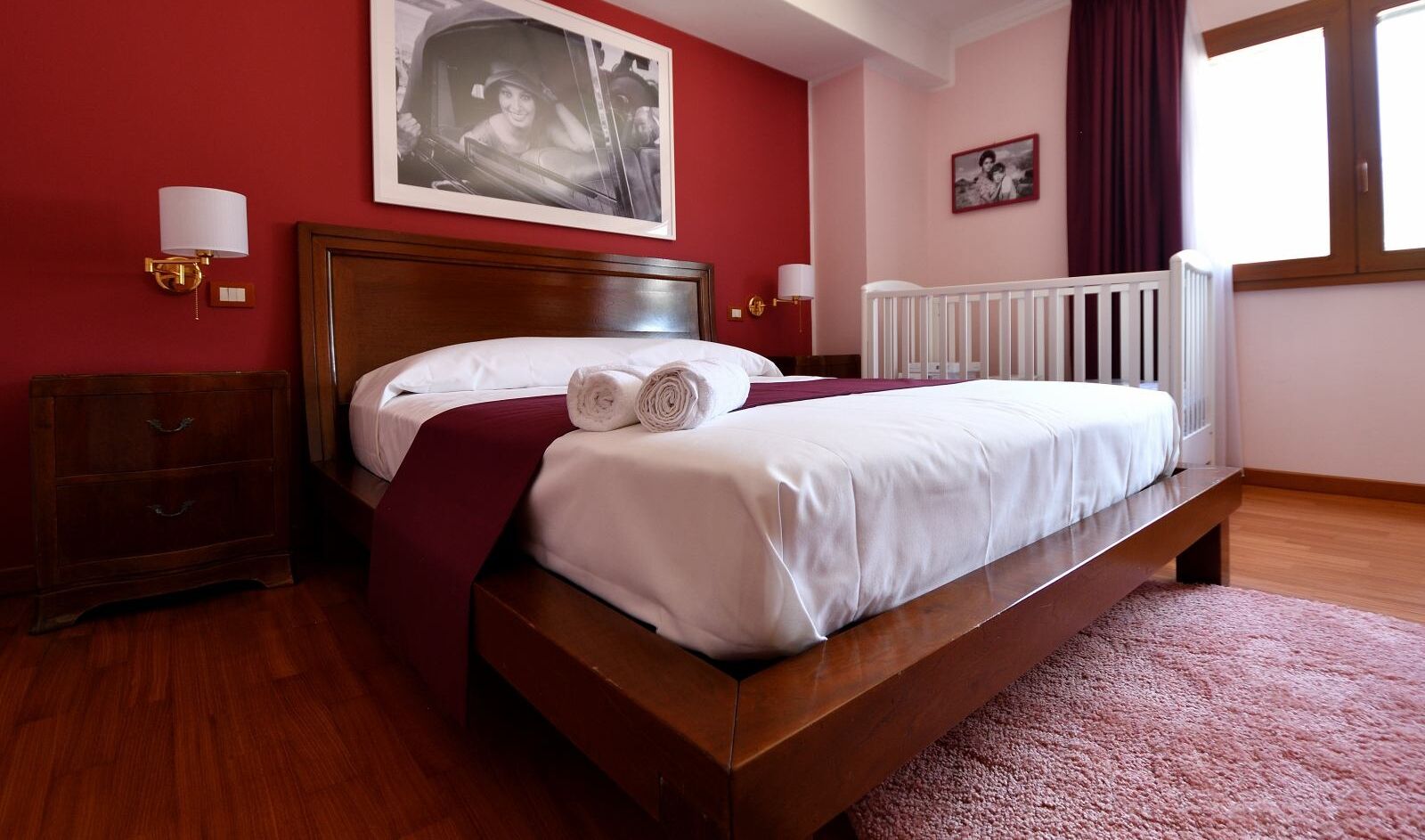 A neatly made bed with white linens and a red throw blanket, a wooden nightstand with a lamp, and a large window with curtains.