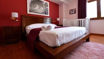 A neatly made bed with white linens and a red throw blanket, a wooden nightstand with a lamp, and a large window with curtains.
