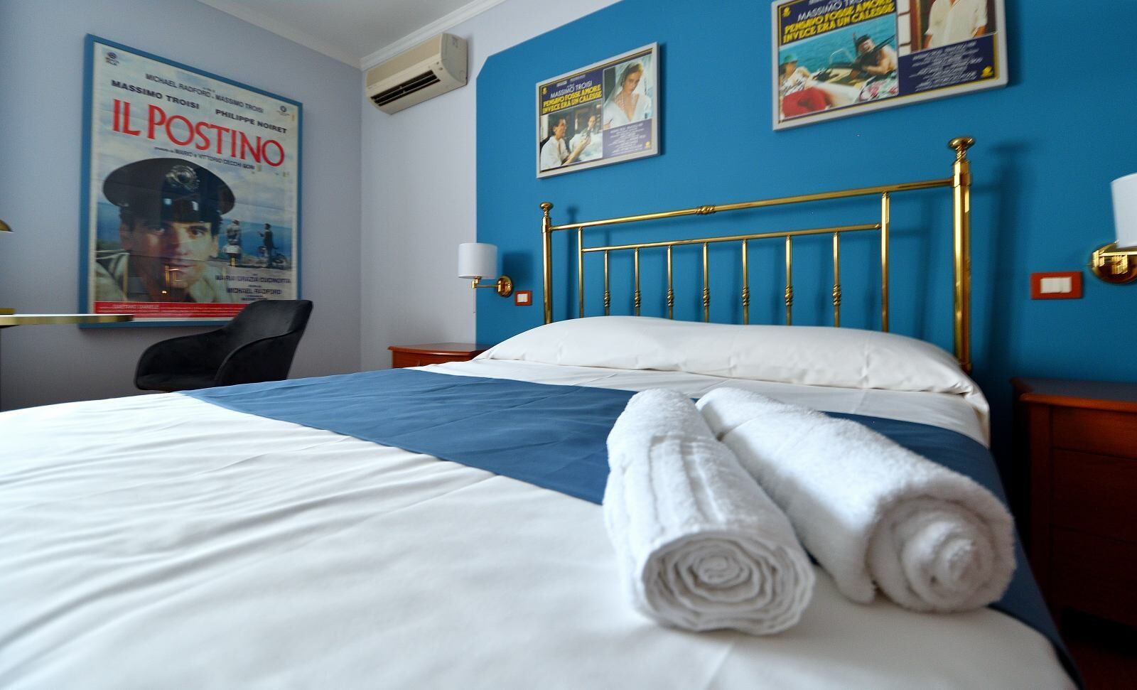 A hotel room with a bed, a desk, and a chair. There are posters on the wall and a window with curtains.