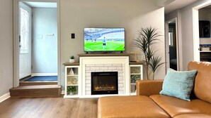 Ein Wohnzimmer mit Kamin, einem Fernseher, der ein Sportspiel zeigt, einem braunen Ledersofa und einer Tür zu einem anderen Raum.