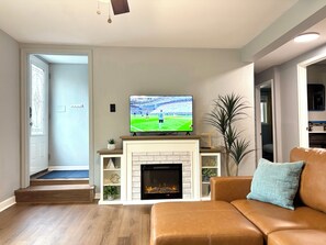 A living room with a fireplace, a television displaying a sports game, a brown leather sofa, and a door leading to another room.