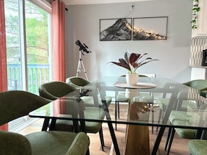 Uma sala de jantar com uma mesa de vidro, cadeiras estofadas verdes, uma planta em vaso e pinturas de paisagens montanhosas na parede.