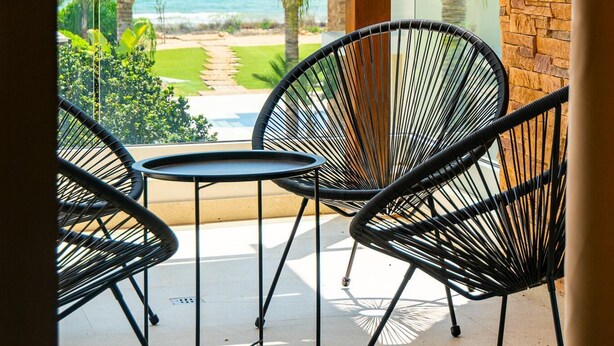 A balcony with a view of the ocean, featuring a glass railing, a wooden column, and black wire chairs.