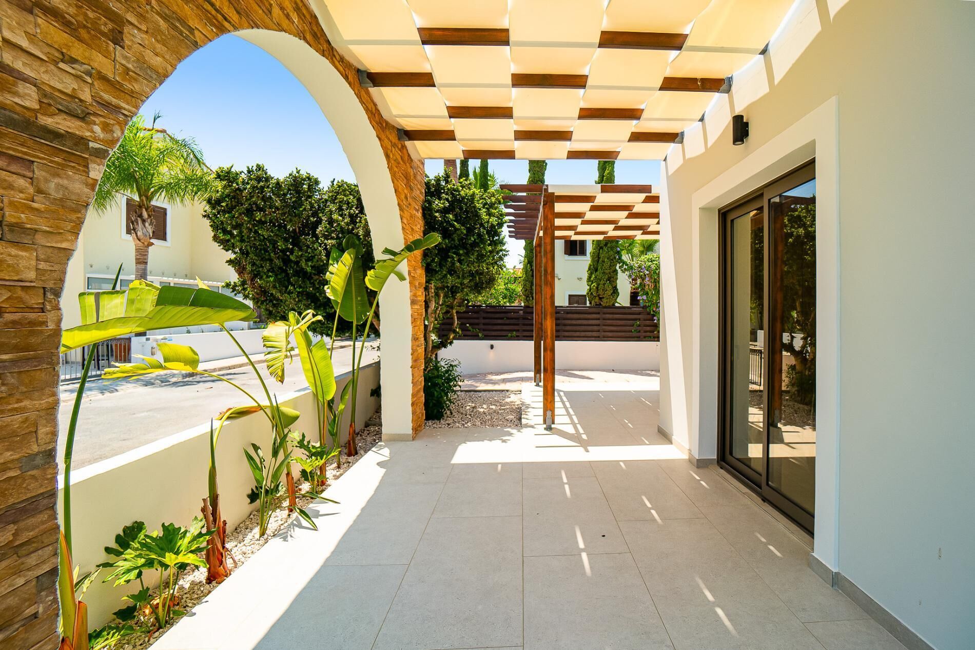 Et overdækket udendørs område med et pergola, beplantning og en sti til en bygning.