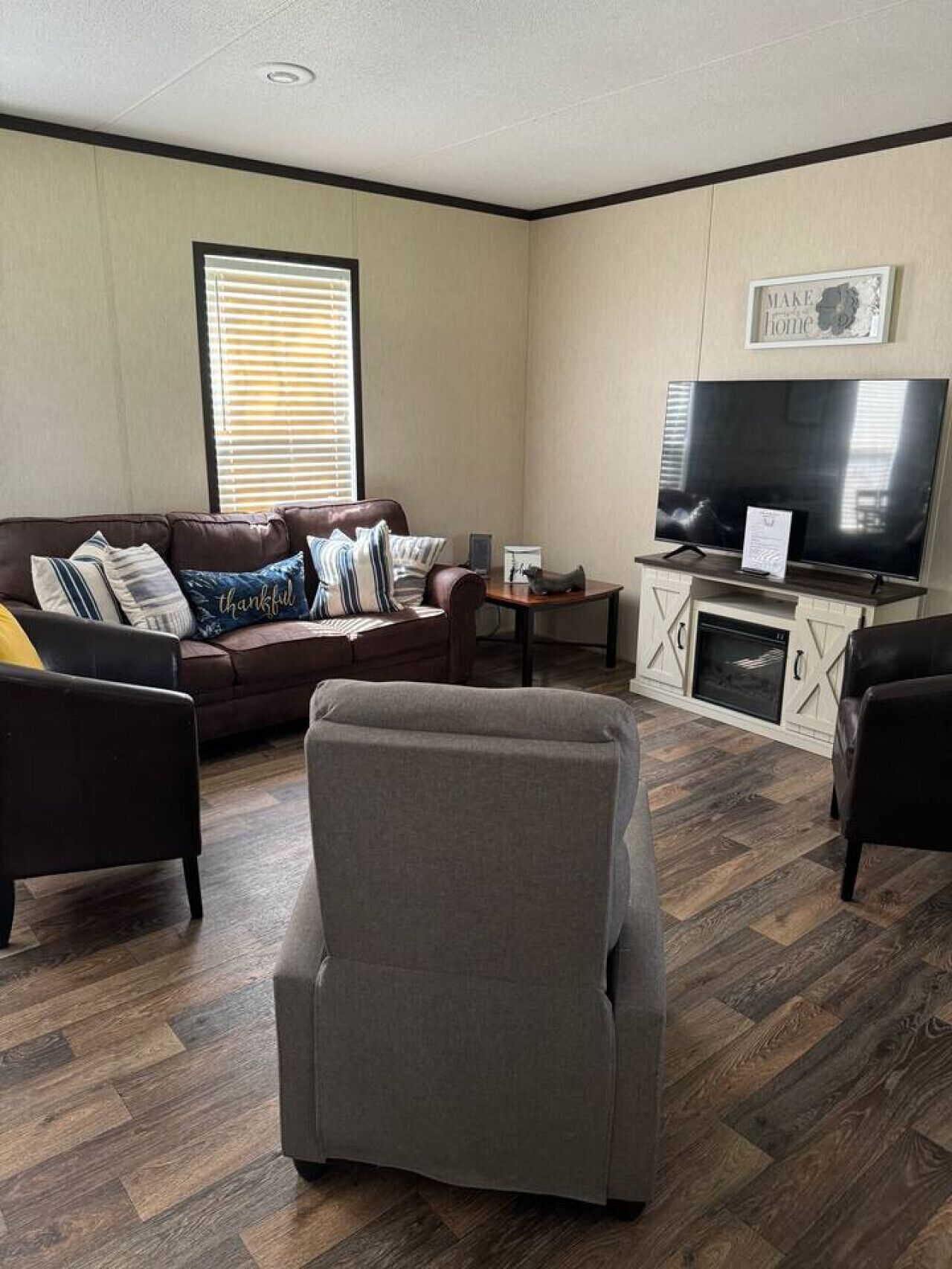 A living room with a brown leather sofa, a grey armchair, a television on a cabinet, and a window with blinds.