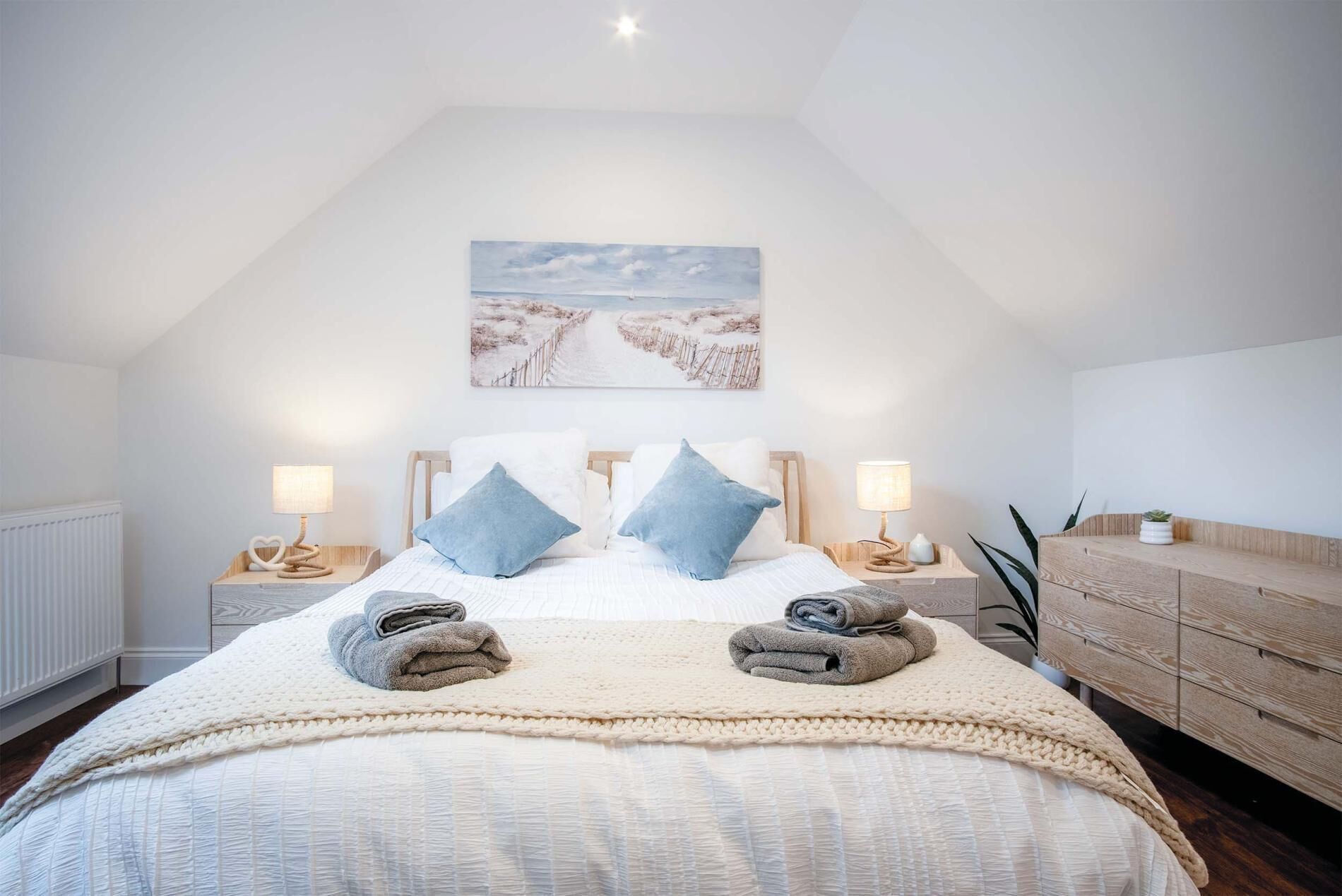 Un dormitorio con techo alto, una cama grande con dos almohadas, una mesita de noche con lámpara y un aparador de madera.