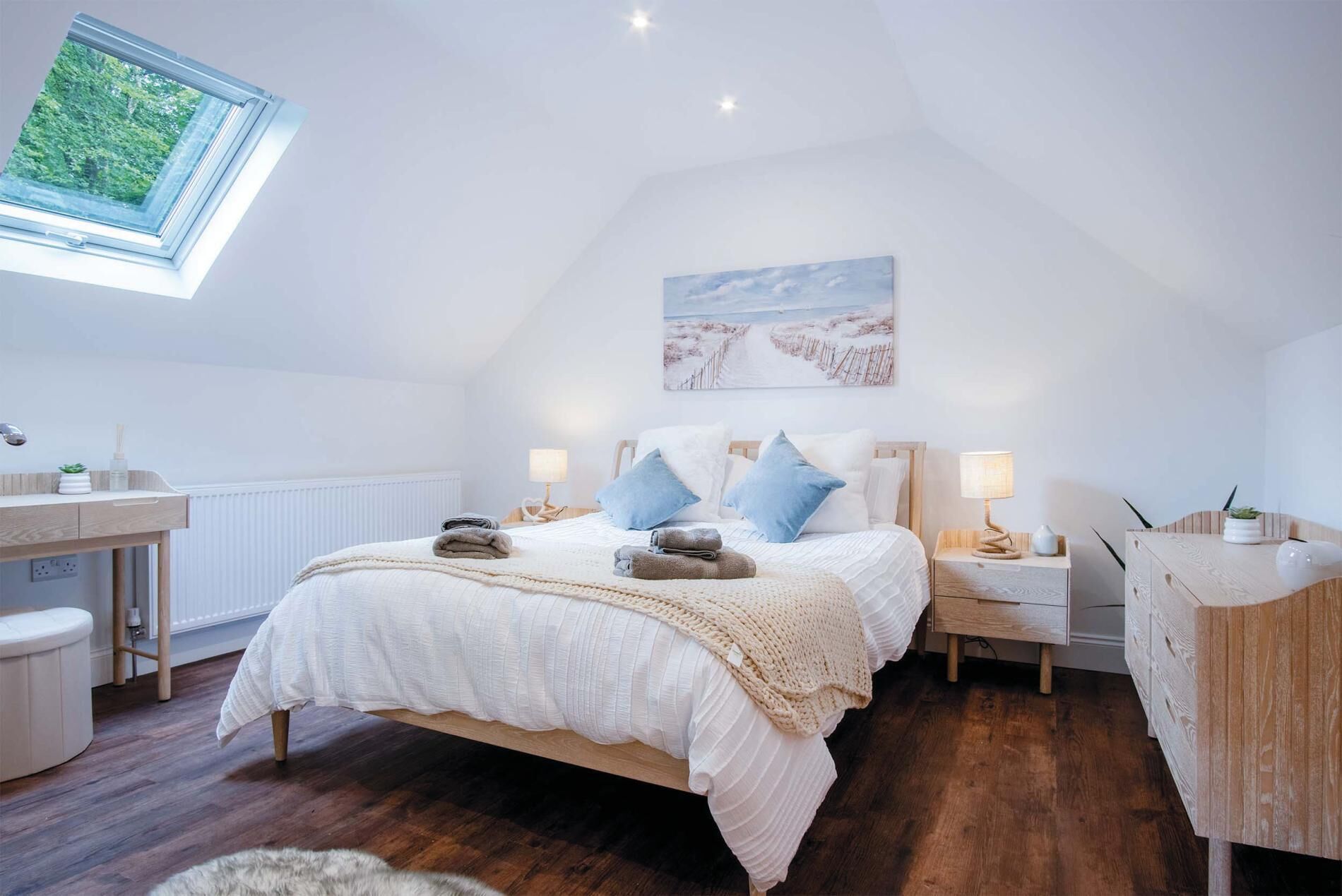 A bedroom with a skylight, a bed with a white duvet and blue pillows, a bedside table with a lamp, and a painting on the wall.