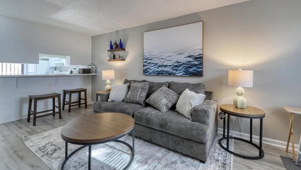 Ein Wohnzimmer mit grauem Sofa, rundem Couchtisch und zwei Beistelltischen inklusive Lampen. An der Wand hängt ein großes Gemälde, das eine Meereslandschaft zeigt.