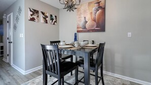 Ein Essbereich mit einem dunklen Holztisch, gedeckt für vier Personen, umgeben von schwarzen Stühlen. An den Wänden hängen abstrakte und florale Gemälde, über dem Tisch hängt eine Chandelier, und an der Wand befindet sich eine große Vase mit einem Gemälde.
