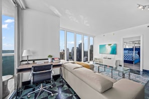 A modern hotel room with a large window, a sofa, a desk with a chair, a TV, and a bed with a view of the city. - Icon Brickell 2BDR 46th Fl Ocean & Pool Views (Miami)