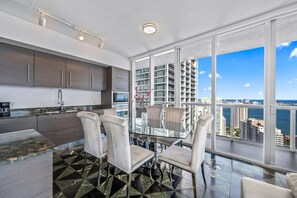 A modern kitchen with dark cabinets, a glass dining table, and chairs, overlooking a cityscape with high-rise buildings.