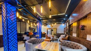 Un interior moderno con una mesa de madera central rodeada por sofás capitonados, iluminación colgante y una pared decorativa con un diseño geométrico.