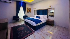 Habitación de hotel con diseño moderno, que incluye una cama grande con ropa de cama azul, una mesita de noche, un espejo e iluminación geométrica en el techo.