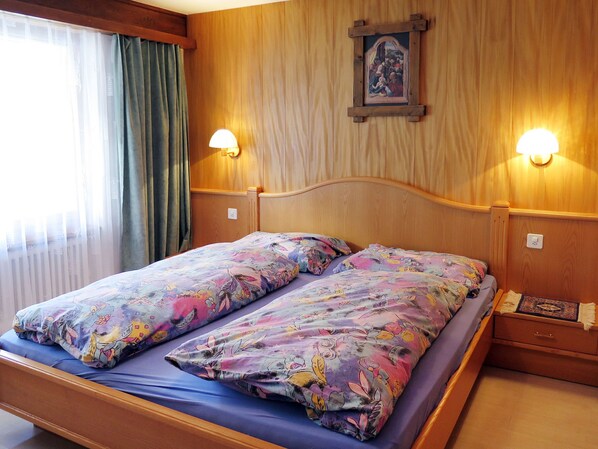 A double bed with floral bedding, wooden headboard, two wall-mounted lamps, and a small bedside table with a decorative item.