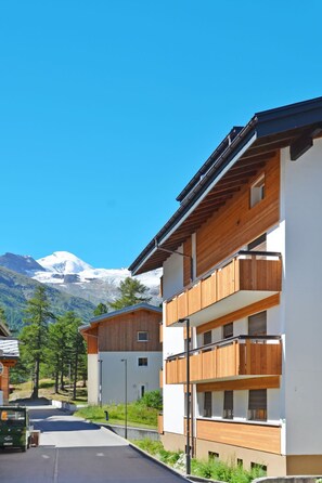 A modern multi-story building with wooden balconies, set against a backdrop of snow-capped mountains and clear blue sky.