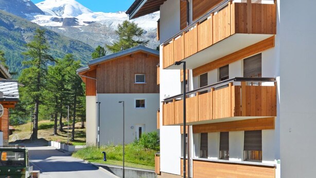 A modern multi-story building with wooden balconies, set against a backdrop of snow-capped mountains and clear blue sky.