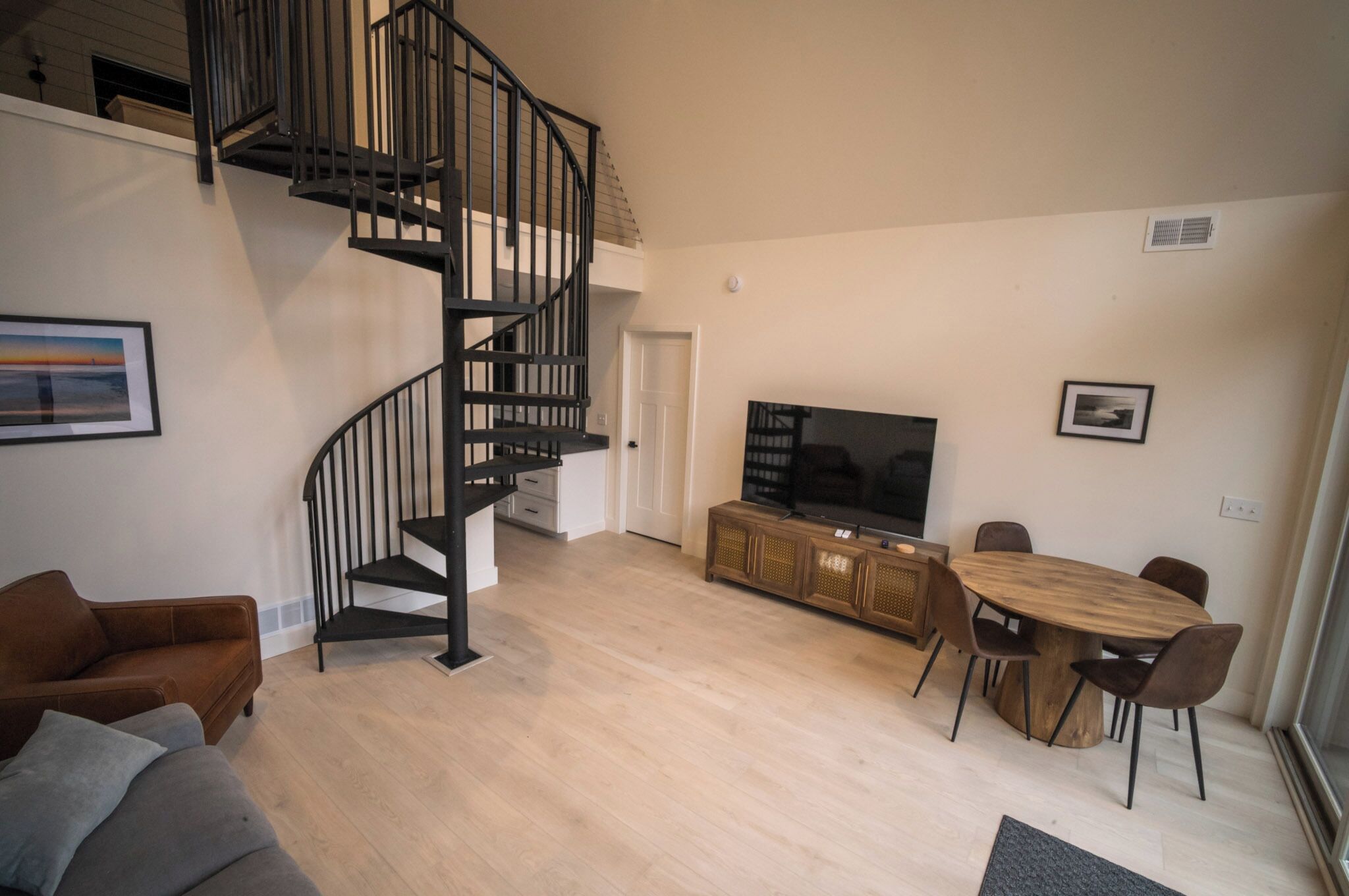 A modern living room with a black spiral staircase, a flat-screen TV, a dining table with chairs, and a sofa.