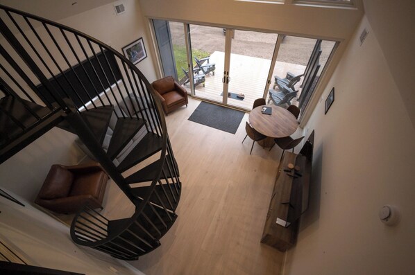 Un interior moderno con una escalera en espiral, una zona de estar con mesa y un gran ventanal corredizo que da a un patio exterior.