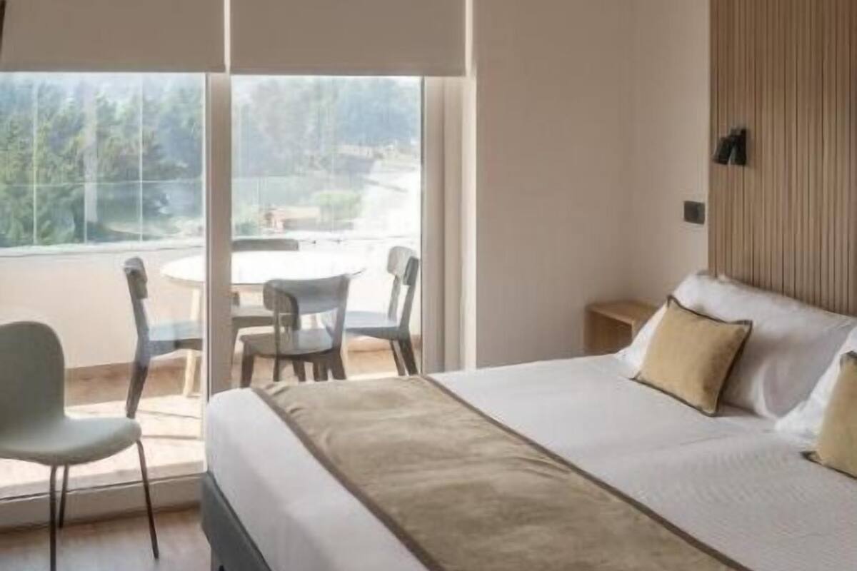 A modern hotel room with a large bed, a small table, two chairs, and a balcony with a view.