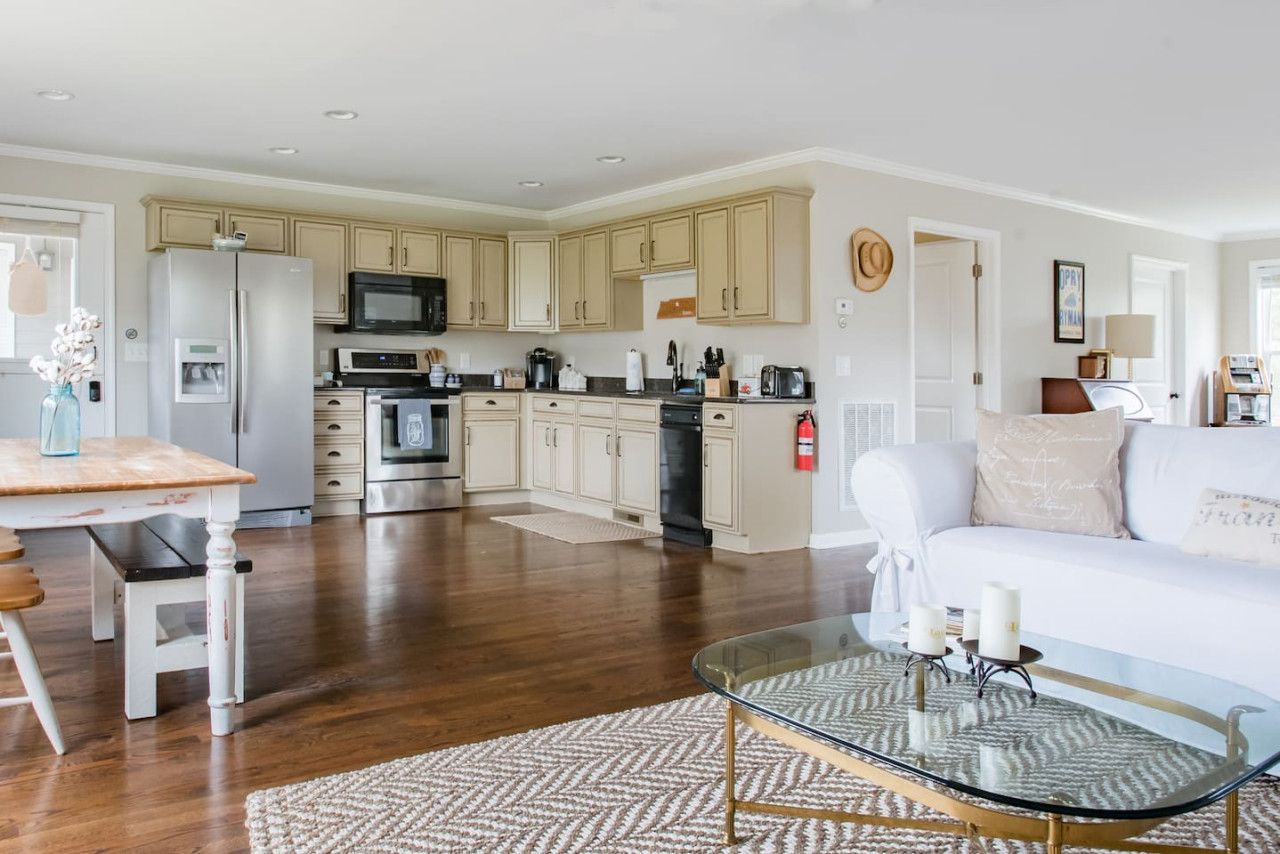A spacious living area with a glass coffee table, a white sofa, and a kitchen in the background.