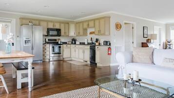A spacious living area with a glass coffee table, a white sofa, and a kitchen in the background.