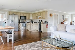 Un amplio salón con una mesa de centro de vidrio, un sofá blanco y una cocina al fondo.