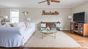 Quarto com cama, mesa de centro de vidro, ventilador de teto, televisão e um letreiro fixado na parede.