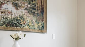 A room with a white wall, a large landscape painting, a white chair, a wooden dresser with a vase of flowers, and a sign that reads 'Be our guest.'