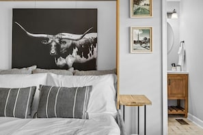 Une chambre à coucher avec un grand lit, une table de chevet, une lampe fixée au mur et des œuvres d’art.