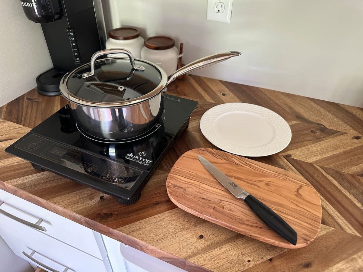 A kitchen counter with a portable induction cooktop, a pot, a white plate, and a wooden cutting board with a knife.