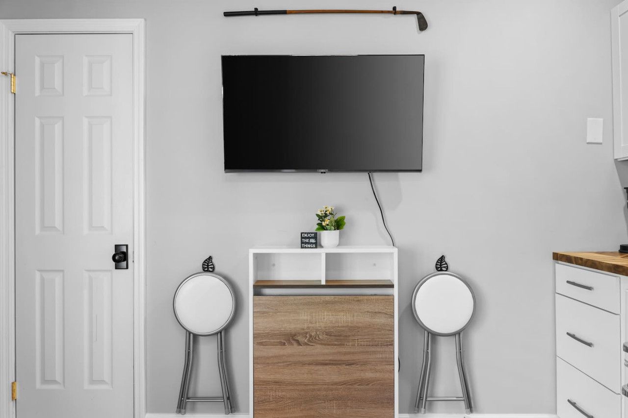 A modern room with a flat-screen TV mounted on the wall, a small wooden cabinet, two white stools, and a white door.