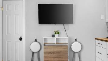 A modern room with a flat-screen TV mounted on the wall, a small wooden cabinet, two white stools, and a white door.