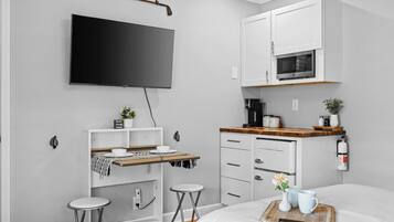 A modern bedroom with a bed, a TV mounted on the wall, a small kitchenette with a microwave, and a bedside table with a tray.