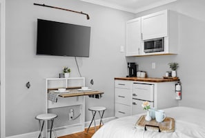 Un dormitorio moderno con una cama, un televisor montado en la pared, una pequeña kitchenette con microondas y una mesita de noche con una bandeja.