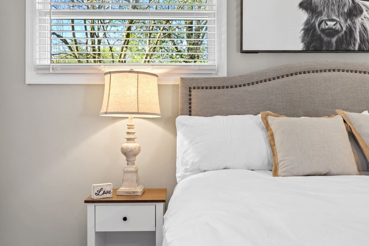A bedroom with a bed, a bedside table with a lamp, a nightstand, and a window with blinds.