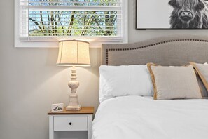 A bedroom with a bed, a bedside table with a lamp, a nightstand, and a window with blinds.