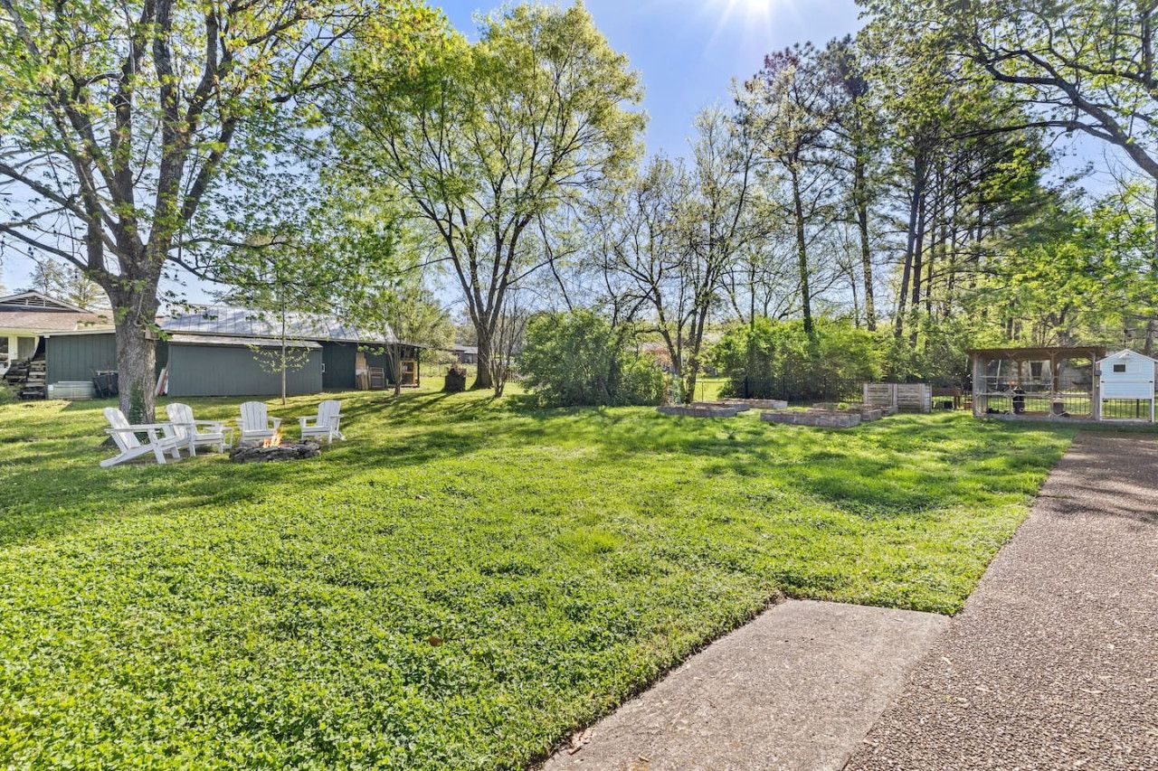 A well-maintained grassy yard with white outdoor furniture and a fire pit, surrounded by trees and a small shed.