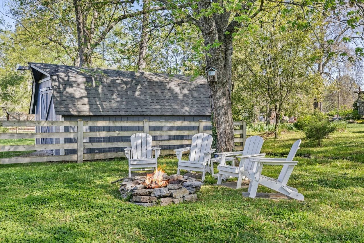 A rustic outdoor seating area with Adirondack chairs and a fire pit, surrounded by a wooden fence and trees.