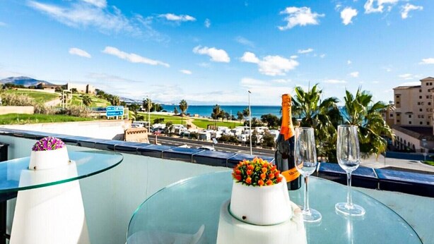 A balcony with a glass table, a bottle of champagne, and two glasses, overlooking a coastal area with buildings and greenery.