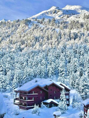 A red building with multiple balconies, surrounded by snow-covered trees and a mountain in the background.