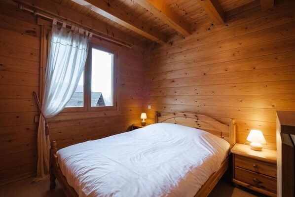 Ein Zimmer einer Holzhütte mit einem Bett, zwei Nachttischlampen, einem Fenster mit Vorhängen und einem Nachttisch aus Holz.