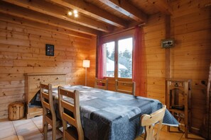 Interior de uma cabana de madeira com uma mesa de jantar, cadeiras, um abajur e uma janela com cortinas.