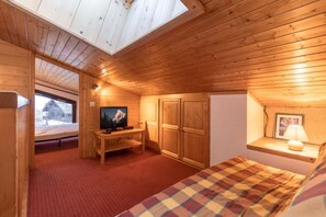 Ein Zimmer in einer Holzhütte mit einem Bett, einem Fernseher, einem Schreibtisch und einem Fenster mit Blick auf eine verschneite Landschaft.