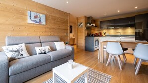 A modern living room with a grey sofa, a small white table, and a kitchen area with a dining table and chairs.