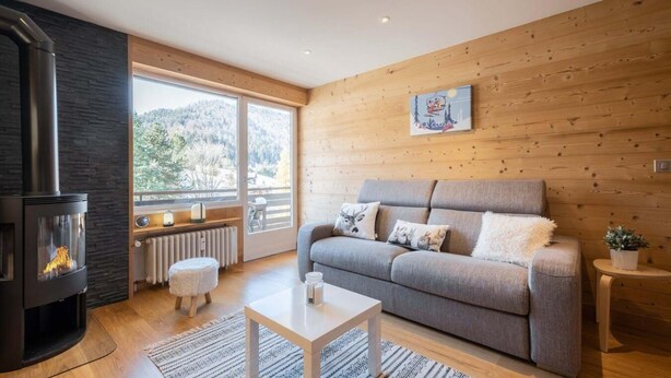 A cozy living room with a fireplace, a grey sofa, a small table, and a view through large windows.