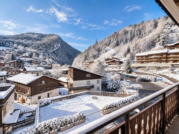 A snow-covered mountain village with chalet-style buildings, a clear sky, and a view of the surrounding forest.