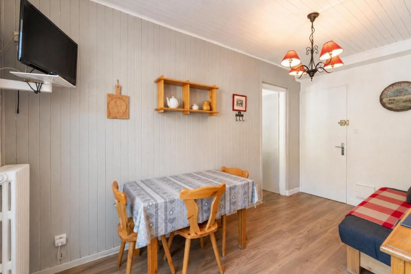 A dining area with a table and chairs, a television mounted on the wall, and a wooden shelf with kitchen items.